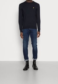 Mann mit schwarzem Pullover mit gelbem Logo, dunkelblauen hochgekrempelten Jeans und schwarzen Leder-Stiefeln bis zum Knöchel, der gegen eine weiße Wand steht.