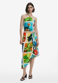 Crop top dengan cetak floral dan kerah halter, dipasangkan dengan rok lilit yang menampilkan pola tanaman cerah dalam panel berwarna-warni. Sandal hitam.