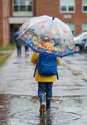 Child in yellow raincoat and blue backpack walks through puddle holding clear umbrella with space-themed designs on a rainy day.