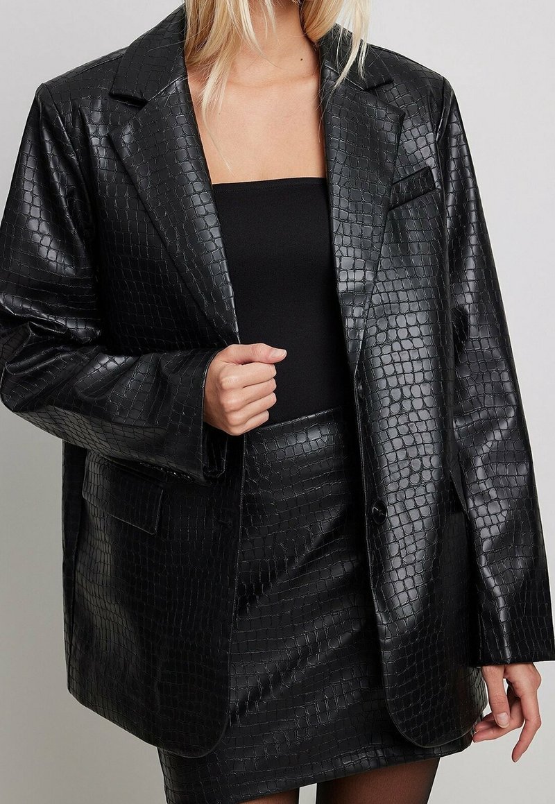 Woman wearing black textured faux leather blazer and matching skirt over a black strapless top against a plain background.