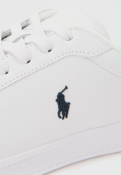 Λευκό δερμάτινο αθλητικό παπούτσι με λευκά κορδόνια και μικρό μαύρο λογότυπο παίκτη πόλο στη πλευρική επιφάνεια κοντά σε ραφές.