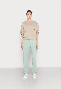Sweatshirt oversized bege com mangas longas, combinado com calças de treino verde-claro. Ténis brancos completam o visual casual contra um fundo simples.
