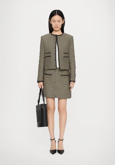 Tweed jacket and skirt set in gold and black with front pockets, gold buttons, and black trim. Accessorized with a black handbag.