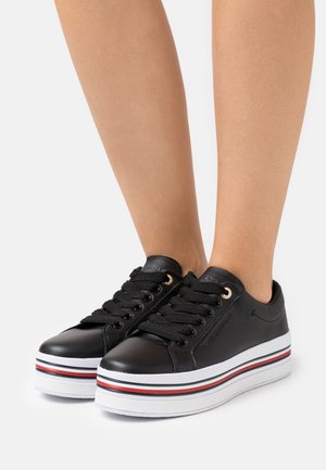 Baskets compensées en cuir noir avec lacets noirs et semelles blanches ornées de bandes rouges et bleues, portées sur jambes nues.