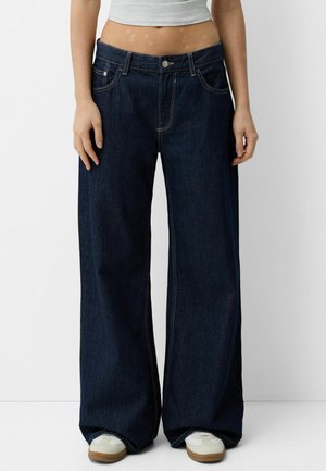 Jeans din denim, de culoare albastru închis, cu croială largă, caracterizate prin buzunare frontale, închidere cu nasture și cusături contrastante galbene. Purtați cu adidași albi.