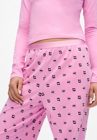 Roze langemouwen top gecombineerd met bijpassende roze broek, voorzien van een herhaald zwart grafisch patroon van gezichten met verschillende accessoires.