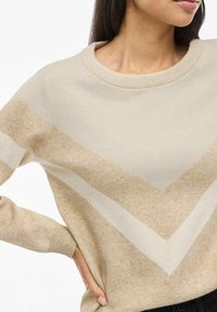 Žena oblečená v béžovom a krémovom svetri s V-vzorom a rebrovaným golierom a manžetami, ruka na bokoch, zamerané od brady po pás.