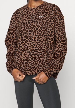 Personne portant un sweat-shirt Nike à manches longues imprimé léopard marron et noir, ainsi qu'un pantalon ajusté gris foncé, mains jointes à la taille.