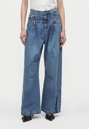 Person iført brede, højtaljede blå jeans med front læg, flere bæltestropper og sorte spidse sko, vist fra taljen og ned.