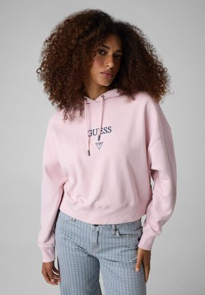 Giovane donna con capelli ricci che indossa una felpa con cappuccio Guess rosa chiaro e jeans a righe azzurre su uno sfondo grigio semplice.