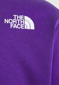 Fialová mikina s žebrovanou texturou, s bílým logem "THE NORTH FACE" na horní levé straně.