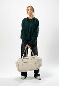 Sac de voyage beige avec double poignées et fermeture éclair. Texture lisse avec des accents latéraux ; modèle portant un sweat-shirt vert et un jean noir.
