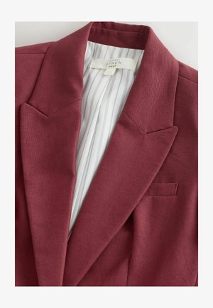 Bordó lenvászon zakó bevágott hajtókákkal és fehér csíkos béléssel, amely egy ruhacímkét mutat a felirattal: „Nyers selyem NEXT.”