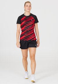 Rød og sort kortærmet træningsskjorte med diagonalt stribemønster, parret med sorte shorts. Model iført hvide sneakers.