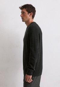 Dunkelgrüner Strickpullover mit strukturierter Oberfläche, breiten Bündchen und Rundhalsausschnitt, kombiniert mit grauen, gemusterten Hosen. Seitenansicht.