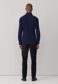 Marineblauwe longsleeve shirt met een kraag, knoopmanchetten en een gladde textuur. Gepaard met donker, slim-fit broeken en bruine schoenen.