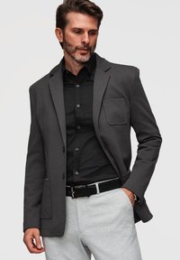 Homme portant un blazer gris foncé, une chemise noire, un pantalon gris clair et une ceinture noire avec la main dans la poche, regardant vers le bas sur le côté.
