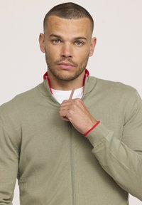 Veste zippée vert olive avec des accents rouges au niveau du col et des poignets, dotée d'une texture lisse et de détails en tricot côtelé.
