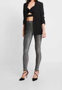 Blazer noir sur un crop top noir tordu, associé à des leggings argentés brillants et des chaussures  à talons noirs. Les leggings présentent une texture pailletée.