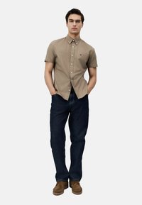 Camisa beige de manga corta con botones y un logo en el pecho, combinada con jeans oscuros y calzado marrón. Diseño simple y casual.