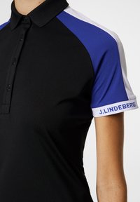 Svart pikétröja med blå och vita raglanärmar, med krage och en logga på ärmen. Slät, atletisk tygstruktur.