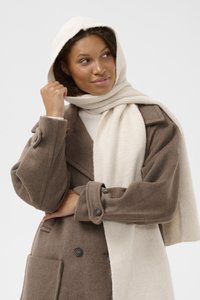 Hnědý vlněný kabát s velkými knoflíky, širokým límcem a texturovanou tkaninou. Krémová šála je volně přehozená kolem krku.