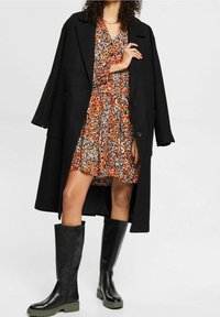 Schwarzer langer Mantel über einem gemusterten Kleid mit oranger Blumenmuster. Knielange schwarze Stiefel mit grünen Sohlen. Glatte Texturen und taillierte Passform.