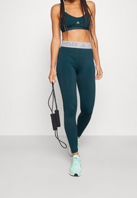 Vihreä urheiluliivi ja korkeavyötäröiset legginsit, joissa on elastinen vyötärö, jossa on harmaa 'ADIDAS' -logo. Yhdistetty mintunvihreisiin urheilukenkiin.