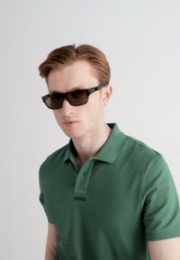 Zelené polo tričko s mírným leskem, s límečkem a logem "BOSS". Černé obdélníkové sluneční brýle s tmavými skly. Jednoduché pozadí.