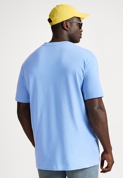 Šviesiai mėlynas medvilninis marškinėlis trumpomis rankovėmis, laisvo kirpimo, su apvalia iškirpte, dėvimas su geltona kepure ir akiniais nuo saulės.