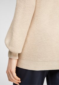 Manche de pull en maille texturée beige avec poignet et ourlet côtelés, portée avec un pantalon foncé, montrant l'avant-bras et le côté du torse.