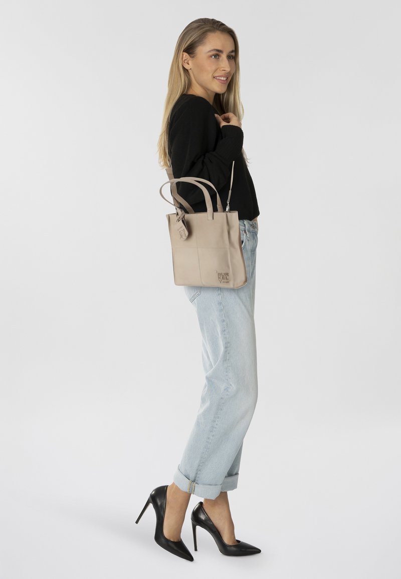 Bolso tote de cuero beige con doble asa y detalle de logo, combinado con jeans de denim azul claro remangados y tacones altos negros.