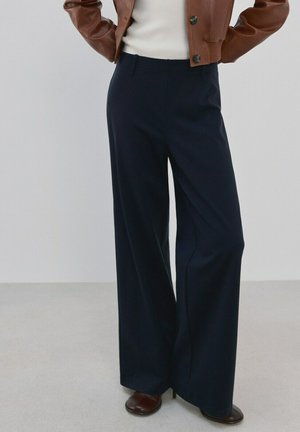 Person iført marineblå bukser med høj talje og vide ben, kombineret med brune hælede sko, en hvid top og en croppet brun læderjakke.