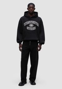 Černá mikina s kapucí a bílým textem "WRSTBHVR STUDIO", doplněná černými kalhotami s bočními zipy a uvolněným střihem. Celkový vzhled doplňují ploché boty.