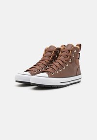 Hnědé kožené vysoké tenisky s bílými gumovými podrážkami, kovovými očky a kontrastními béžovými akcenty na jazyku a límci.