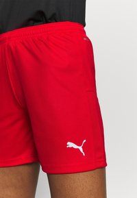 Pantalones cortos deportivos rojos hechos de una tela ligera, con un panel de malla en el lateral y un logo blanco en la parte inferior de la pierna izquierda. Incluye cinturilla elástica.