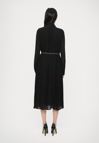 Γυναίκα με την πλάτη γυρισμένη φοράει ένα μαύρο πλισέ midi φόρεμα με μακριά μανίκια, μια ασημένια ζωνάρα και μαύρες ψηλές γόβες.