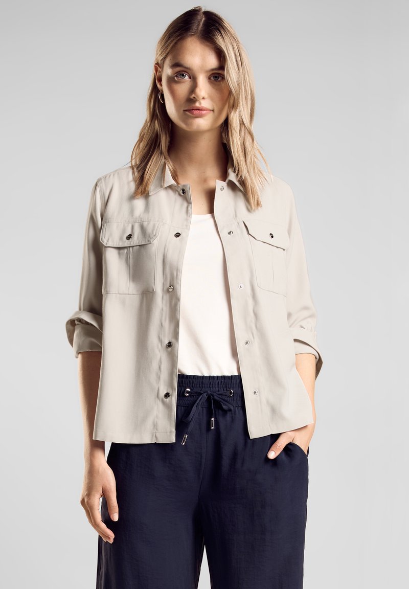 Vaaleanbeige pitkähihaisessa nappipaidassa on kaksi rintataskua ja käännetyt hihat, jotka on yhdistetty tummansinisiin, narukiristettävien vyötäröiden housuihin. Paita on sileäpintaista materiaalia.