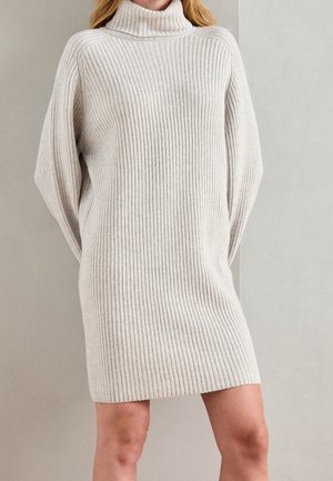 Woman wearing a cream-colored, ribbed knit turtleneck sweater dress with long sleeves standing against a plain background.
