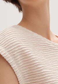 Texturiertes, hellbeiges Gewebe mit Wellenmustern und einem subtilen Glanz, das über einen weiten, abgerundeten Ausschnitt und kurze Ärmel für eine lässige Passform verfügt.