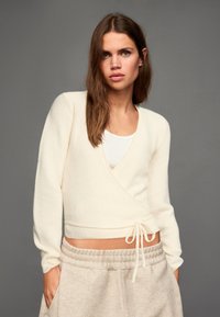 Džemper u krem boji od mekanog materijala s dugim rukavima, dubokim izrezom i strukom s vezicom, uparen s bež trenirkom.