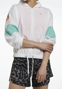 Donna che indossa una giacca bianca con zip, con accenti turchesi e corallo, e pantaloni corti sportivi neri e grigi a fantasia, mentre sistemano la manica del braccio sinistro.