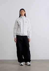 Λευκό μπουφάν με φερμουάρ και μαύρες λεπτομέρειες, σε συνδυασμό με φαρδιά μαύρη παντελόνια cargo. Το μοντέλο φοράει λευκά αθλητικά παπούτσια με μαύρες λεπτομέρειες. Φόντο τσιμεντένιο δάπεδο.