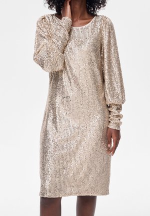 Mujer con un vestido dorado de lentejuelas de manga larga y longitud hasta la rodilla, con escote redondo, posando frente a un fondo blanco.