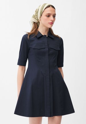 Kobieta w granatowej sukience z kołnierzykiem, z półdługimi rękawami i kieszeniami z patką, nosząca kremowo-zielony pasiasty szalik na głowie.