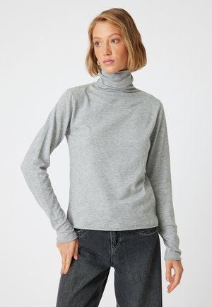 Un col roulé gris en tissu doux avec des manches longues, présentant une coupe ajustée et une texture lisse. Porté avec un jean foncé.