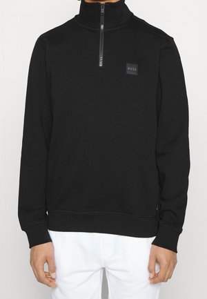 Zwarte longsleeve trui met kwartritssluiting en BOSS-logo op de borst, gedragen door een persoon met witte broek, staand tegen een effen achtergrond.