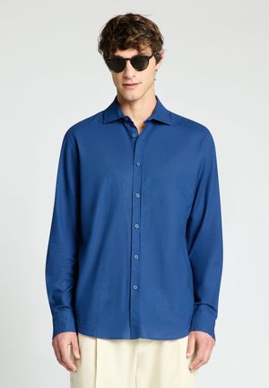 Junger Mann mit schwarzen Sonnenbrillen, einem blauen Hemd mit langen Ärmeln und hellen Hosen steht vor einem einfarbigen Hintergrund.