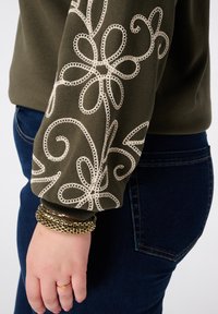 Groene sweatshirt met ingewikkelde witte bloemembroidery op de mouw. Gecombineerd met donkerblauwe jeans en meerdere gouden armbanden.