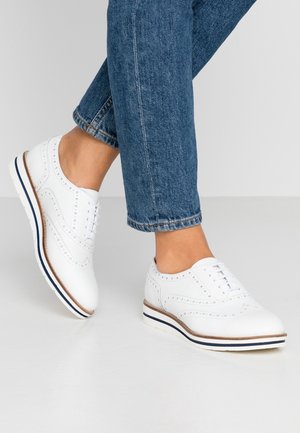 Person iført hvide læder brogue-sko med blå og brune stribede såler, kombineret med blå denimjeans, der er croppede ved anklen.